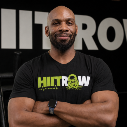 A smiling man with a beard stands with arms crossed, wearing a black HIITROW club fit t-shirt in front of a wall with large HIITROW text.