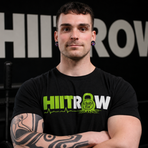 A person with short dark hair, tattoos, and piercings stands confidently with arms crossed, wearing a black HIITROW club fit shirt in a gym setting.