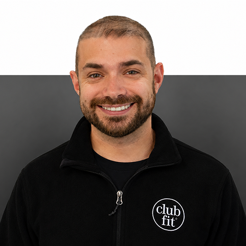 Man with short brown hair smiles infront of half grey half white background, wearing club fit jacket.